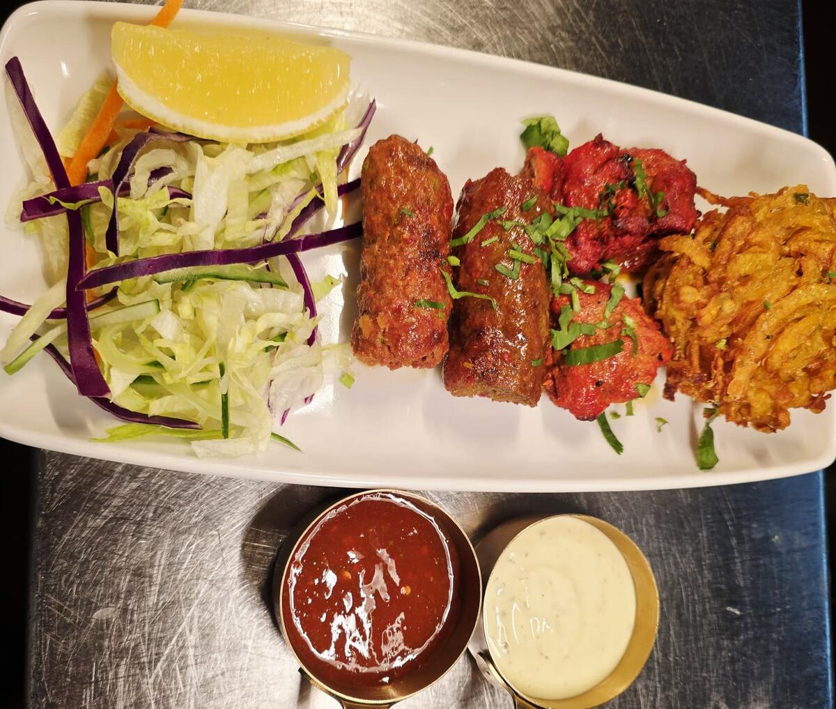 A tandoori mixed grill platter seen from above: seekh kebab, tandoori chicken and onion bhaji with salad, lemon and two dips