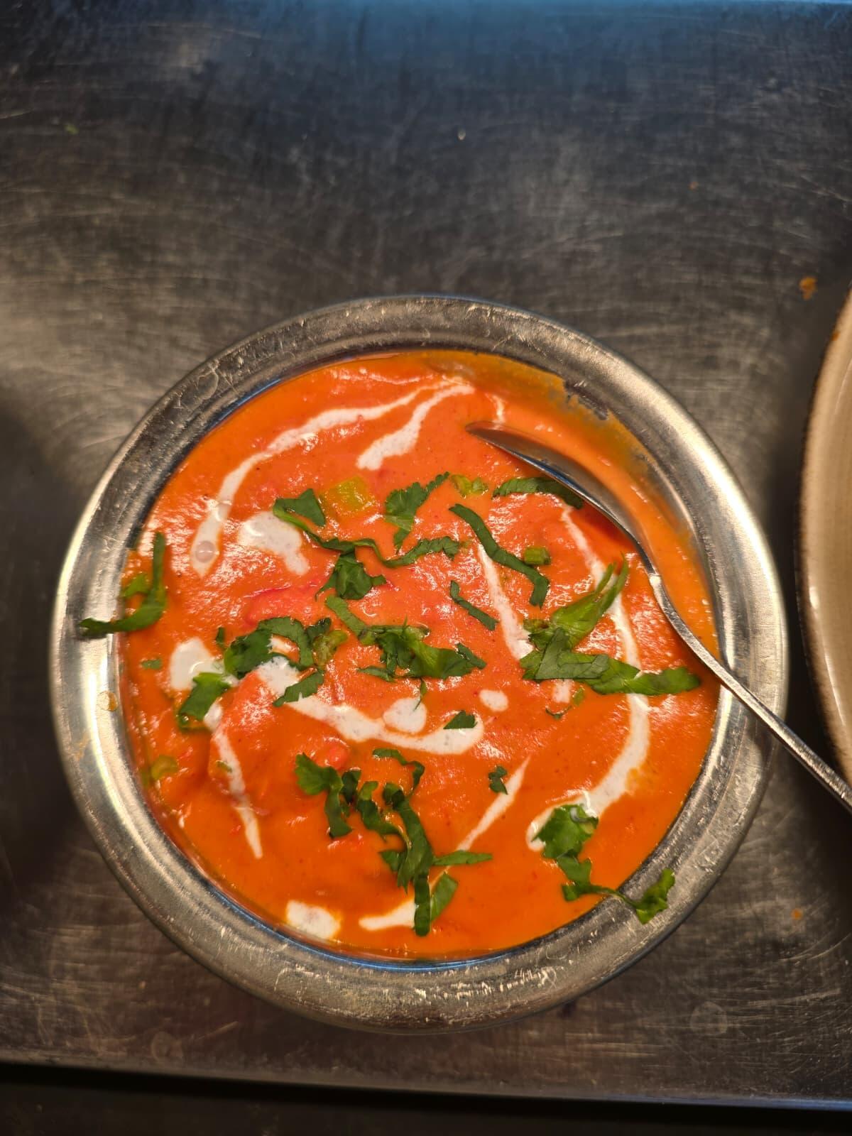 A metal bowl of creamy tomato curry with a swirl of cream and chopped coriander